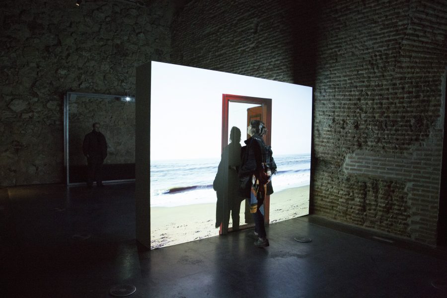 person in a dark room looks at a bright screen with an image of a beach and crashing waves. In front of the breach is an open red door.