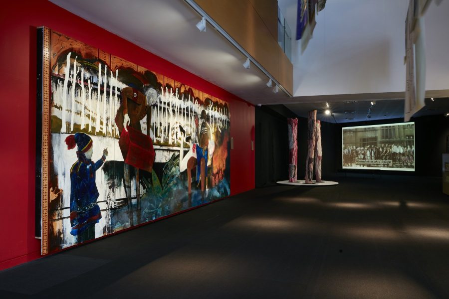An expansive gallery space. A large mural hangs on the left red wall. Banners hang from the ceiling, and a sculptural form is pictured in the background.
