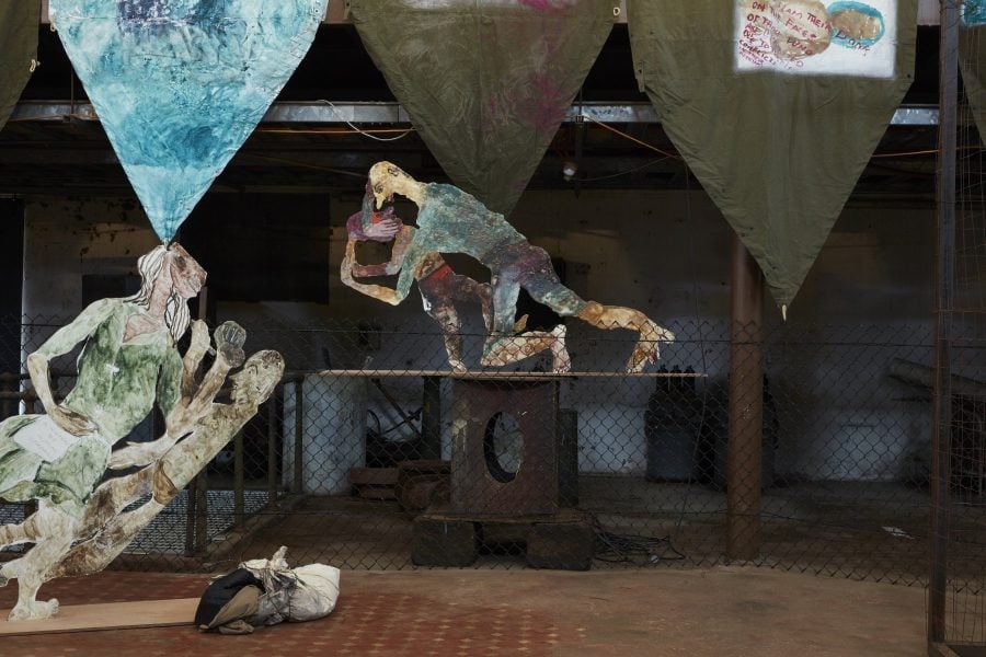 Painted wooden figures are assembled in a large industrial space. Painted green banners also hang in the space.