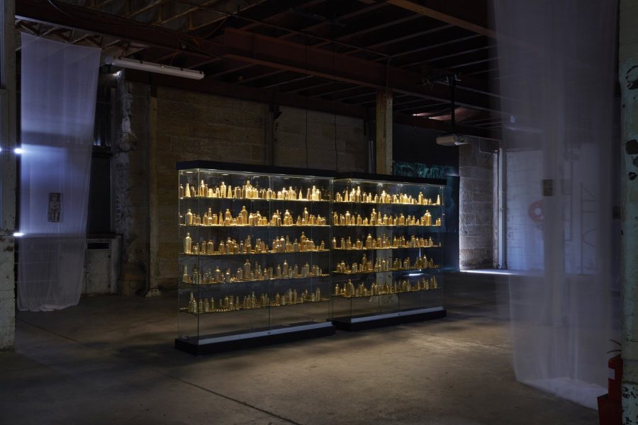 A glass cabinet approximately 4 meters long stands packed with golden bottles. In the space is a number of thin white fabric pieces hanging from the ceiling.