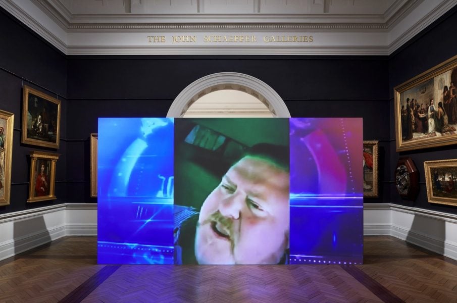 A large screen stands in the middle of a traditional gallery space. Paintings hand on dark painted walls surrounding the screen. On the screen, a film plays with a close up image of a white man who is talking.