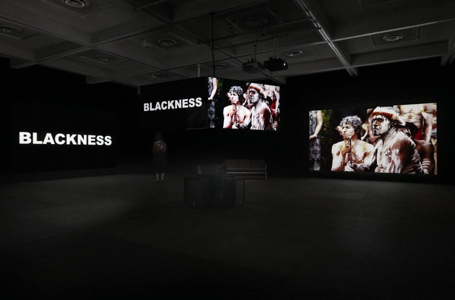 Multiple screens are projected in a dark space. Screens suspended from the centre of the room and on the side walls. A photograph of two people with white paint on their bodies is projected in the space, as well as the word