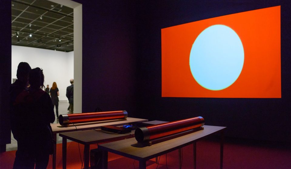 Installation view of a dark gallery room with a projection on the wall of a white circle on an orange/red background.