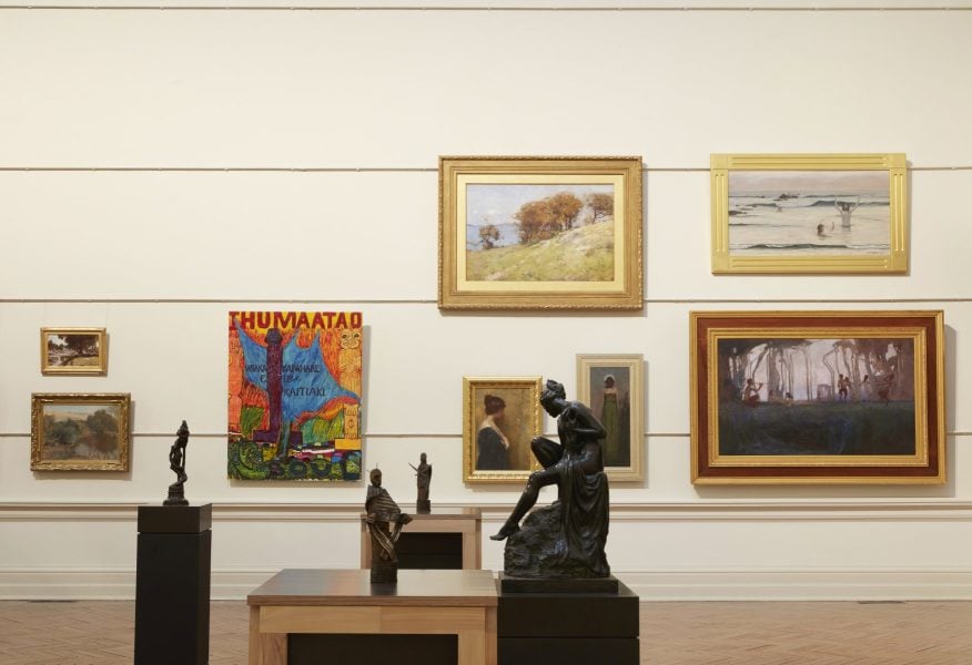 A traditional art gallery space. Various classical landscape paintings are displayed on the back wall, alongside a brightly coloured abstract painting. There are various small metal sculptures on plinths in the middle of the room.
