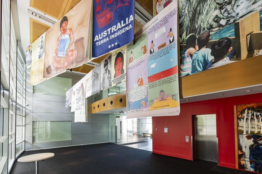 Several different size and colourful posters are suspended from the ceiling in an art gallery foyer. There are two rows of posters. There is a red wall behind and a glass door adjacent. In the left side of the image there is a wall of glass windows.
