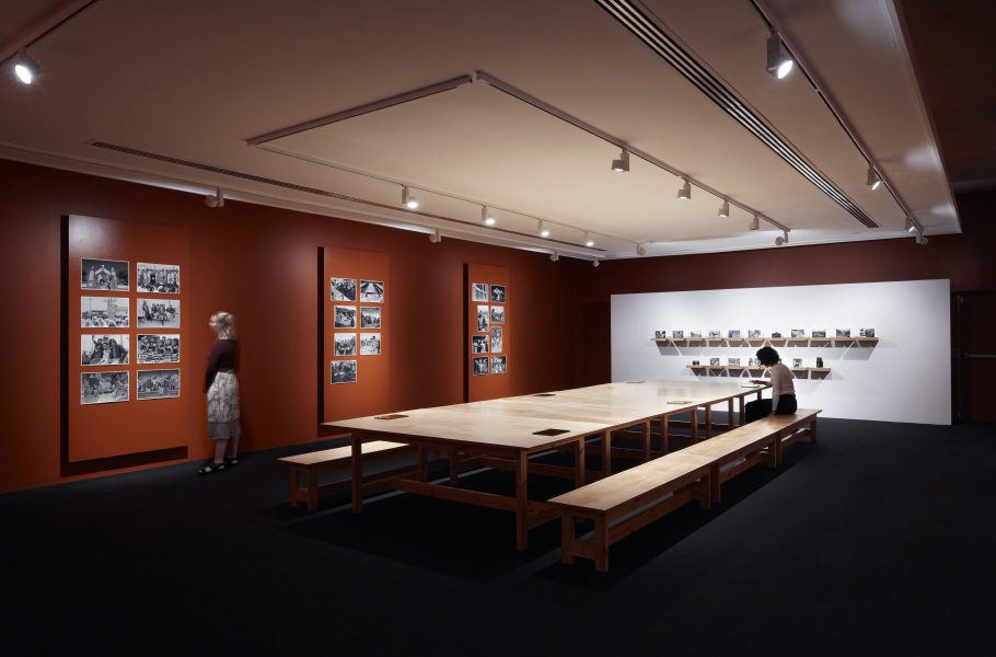 A large space with red walls. Black and white photographs are displayed on the walls. There's a large wooden table in the middle of the room with people sitting at them looking at iPads.