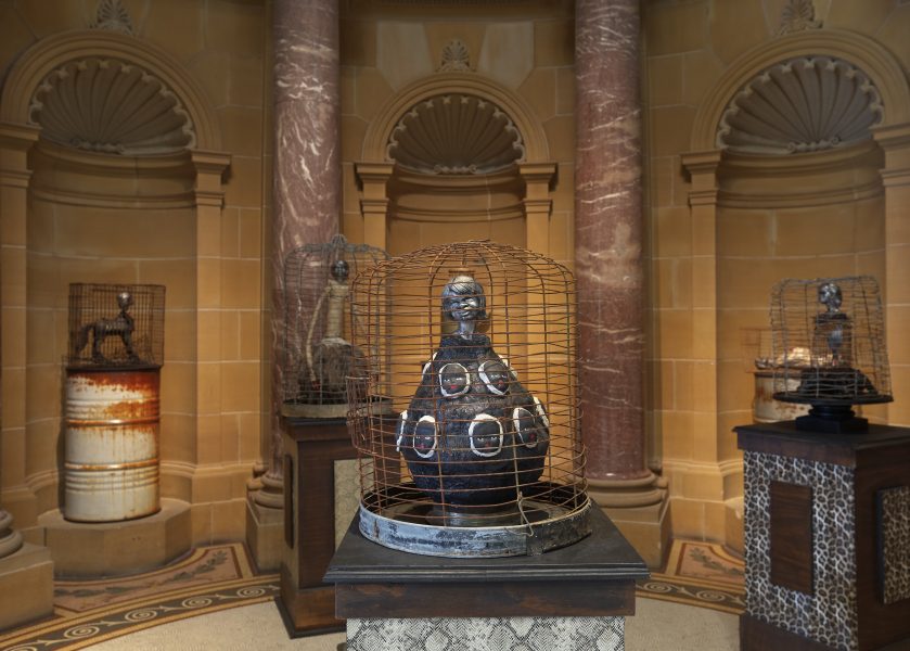 A traditional gallery space with tall stone walls and stone pylons line the space. Small caged metal sculptures sit stop brown plinths.