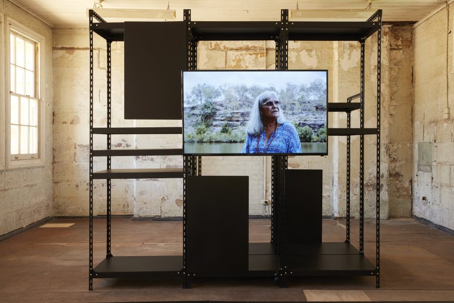Black archive shelves stand in a disused room on Cockatoo Island. A TV hangs on the shelves with a film of Chrissy Doherty speaking about repatriation.
