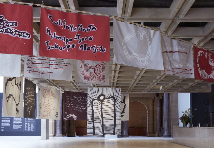 Abstract black and white paintings are suspended from the ceiling of an expansive gallery space. The painting in the foreground is an outline of the Australian continent.