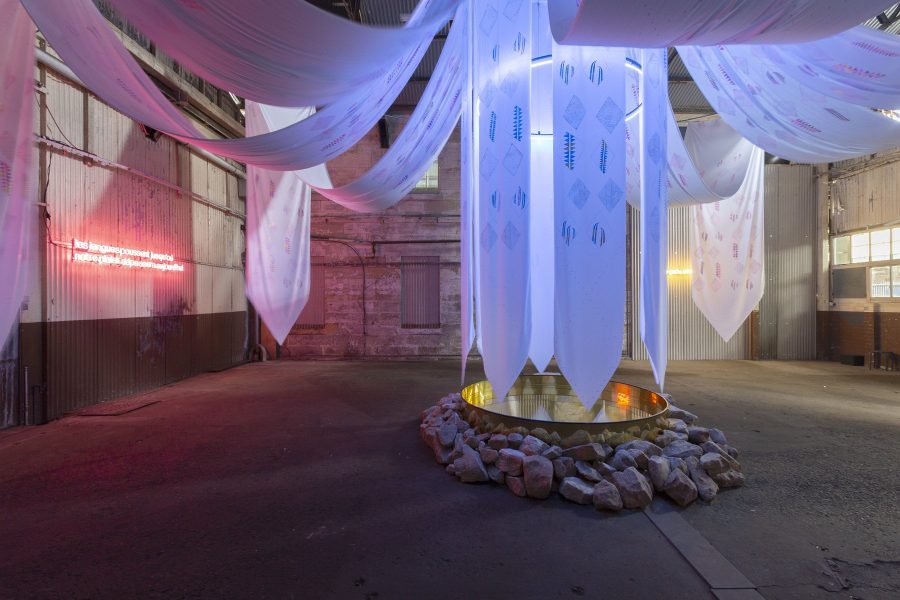 A circular golden water pool sits in the centre of an industrial space with a border of sandstone blocks. White banners with printed motifs are suspended above the pool, extending to the perimeters of the space. A screen and neon phrases are on the walls