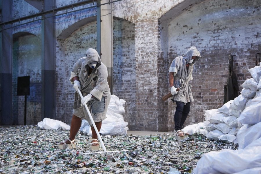 Latai Taumoepeau and Taliu Aloua are standing on a bed of broken glass in a large industrial space. They are wearing grey robes, protective masks and goggles, sandals made of bricks, and white gloves. Latai is raking the glass.