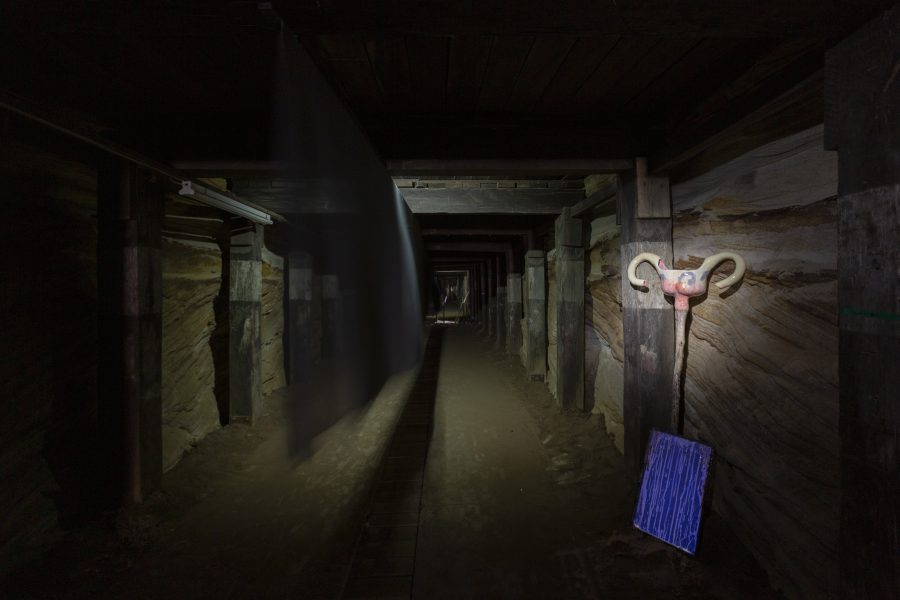 A long dark tunnel with a black curtain hanging in the middle of the space. A TV screen is on the right hand side of the tunnel resting up against the stone wall.