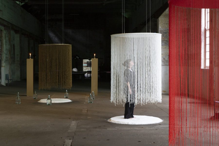 3 long circular veils hang from the ceiling. One gold, one white and one red. Under the veils is a circle of white salt. A person is standing under one of the veils, looking towards the ceiling.