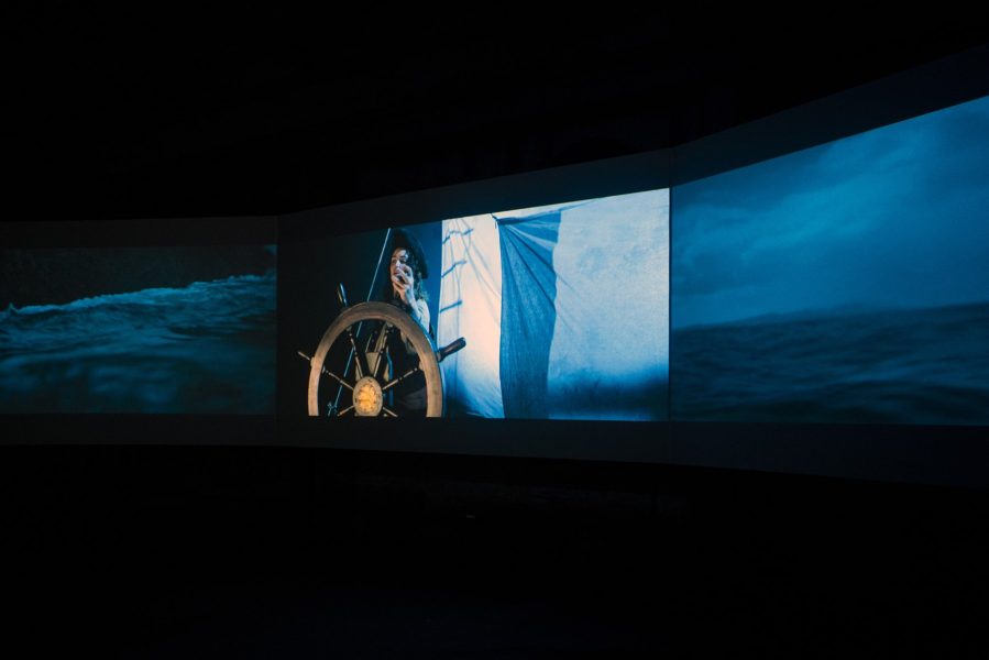 Three screens are suspended side-by-side in a dark space. The middle screen depicts a woman on a boat, standing at the wheel. The other who screens depict the ocean.