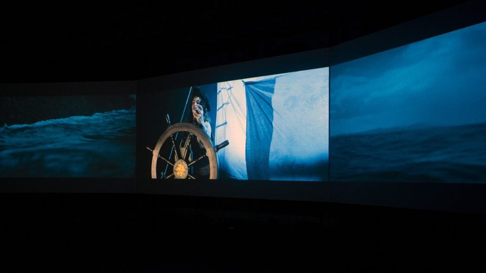 Three screens are suspended side-by-side in a dark space. The middle screen depicts a woman on a boat, standing at the wheel. The other who screens depict the ocean.