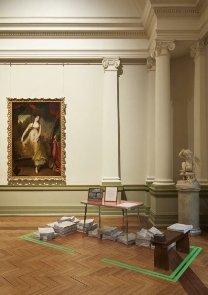 A traditional gallery space with tall white walls. There is a classical oil painting of a woman in a dress on the left. On the right a classical marble sculpture of a baby angel. Centre are many piles of newspaper and a red table over the top of them.