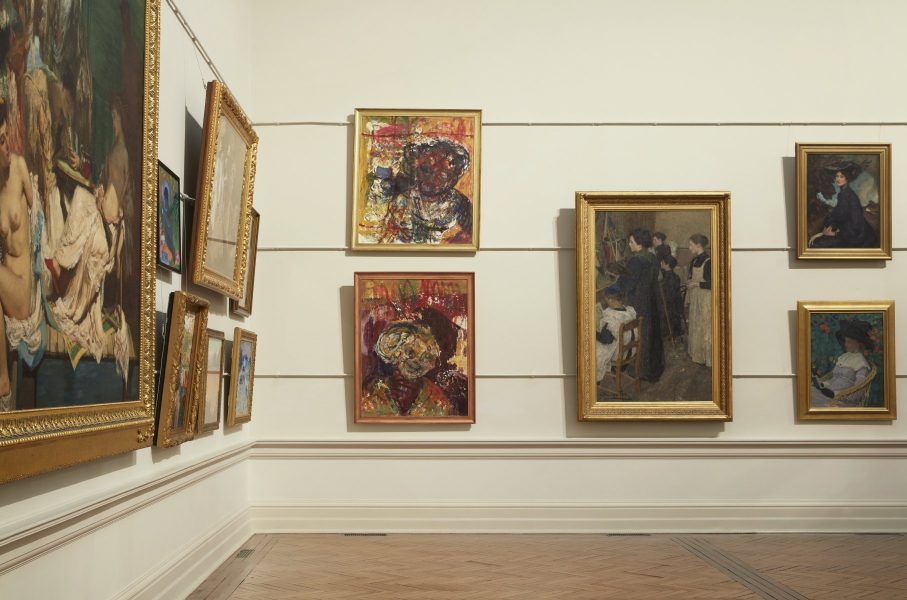 Two paintings by the artist Mostaff Muchawaya hang on a gallery wall amongst collection works at the Art Gallery of New South Wales. The portraits contain abstracted faces and vivid colour markings contrast strongly with surrounding traditional portraits.