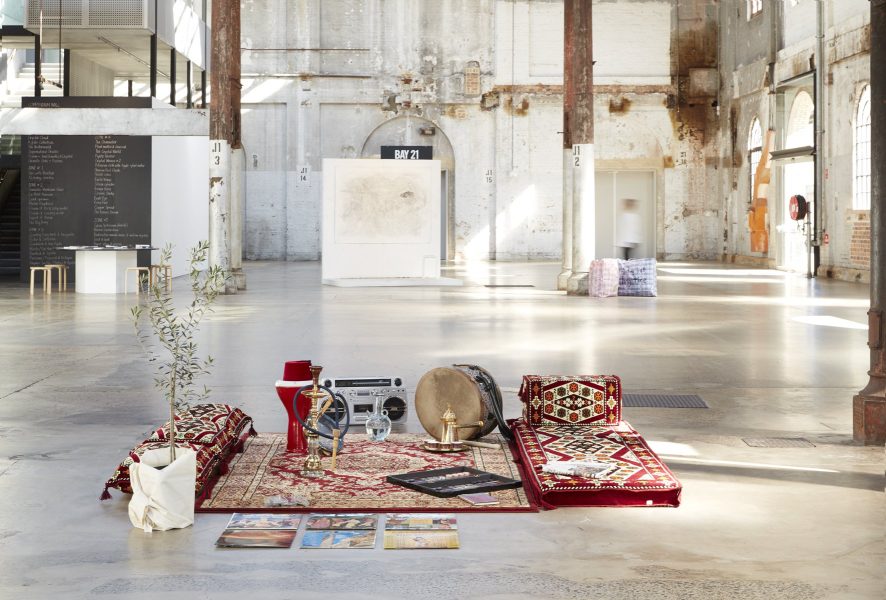 A red Persian rug is on a concrete floor. A small tree is in a white pot next to the carpet. 6 vinyl records are laid out next to the rug.