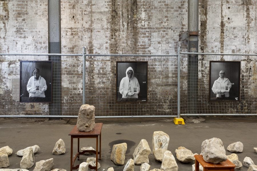 Three framed black and white portrait photographs of women are mounted on a metal fence in front of a brick wall. On the ground in front of them are sandstone blocks with drawings of skulls on them