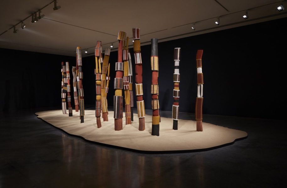 Installation of ironwood tutini poles painted with designs in white, yellow, red and black. The tutini are positioned closely together on a bed of sand in a darkened space.