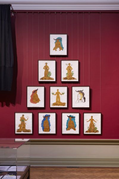 10 framed illustrations are hanging on the dark red wall of a traditional gallery space in a triangle shape. A black curtain hangs to the left of the illustrations.