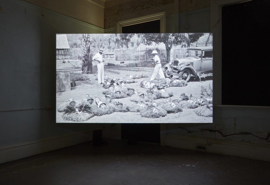 This image depicts a projection of Sammy Baloji's film on a large suspended screen in the corner of a dark room. The scene depicted features two men dressed in white inspecting African artefacts which are splayed across the ground before them.