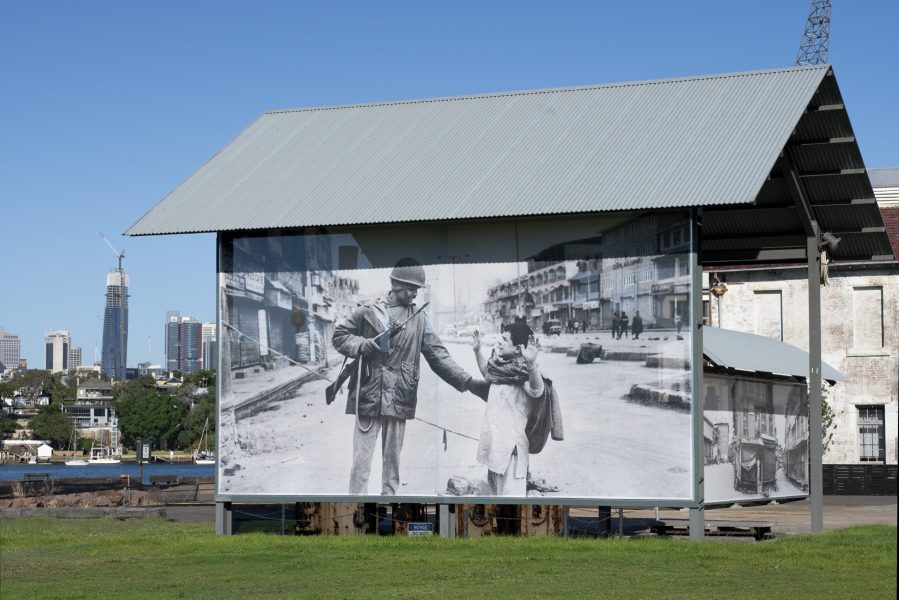 The photo shows a largescale photographic banner by Kashmir / Shaheed / Witness. The photo is black and white and shows a soldier shaking hands with a child in a Kashmir street.