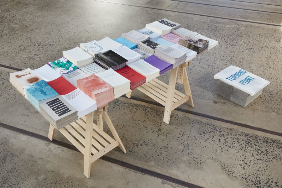 33 piles of papers are assembled in 3 rows on a table. The papers are various colours and one has he word