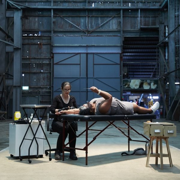 This image depicts a women being tattooed by a traditional tatooist. The tattooist is using thin bamboo to form a temporary mark on the women's arm. Both women are situated side by side in an expansive industrial space.