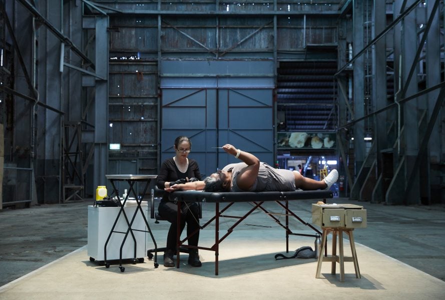 This image depicts a women being tattooed by a traditional tatooist. The tattooist is using thin bamboo to form a temporary mark on the women's arm. Both women are situated side by side in an expansive industrial space.