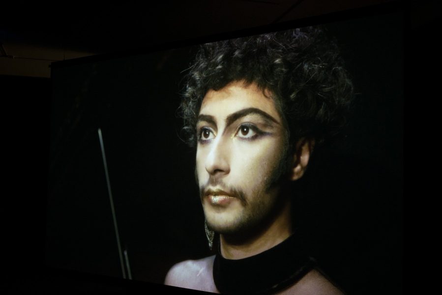 A large screen is suspended on a diagonal in a dark space showing a frame from the video work 'Out of the Blue' by Tarek Lakhrissi