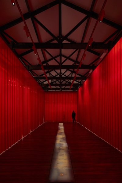 A curtain wall of red butchers curtains surround a line of hot iron plates which lie in the centre of the room. Small puddles of water can be seen in the centre of each hot plate where water has fallen and extinguished.