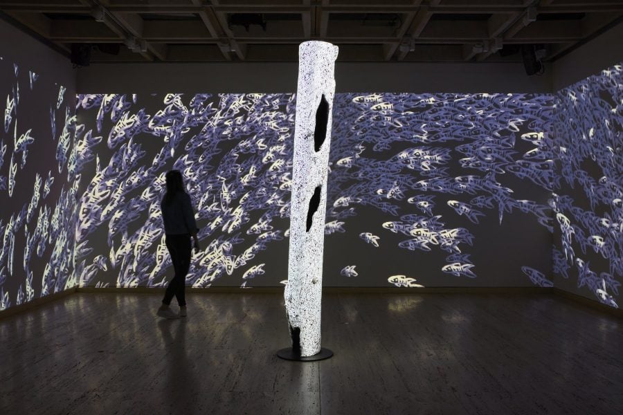 A room has full-size projections on each wall. The projections depict a sea of animated white and purple fish. The silhouette of a person is walking through the space.