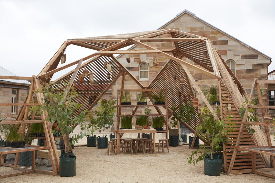 A large wooden octagonal structure sits within the courtyard surrounded by stone convict buildings. There are plants growing in the space. There's a wooden table in the middle of the space with wooden stools around it.
