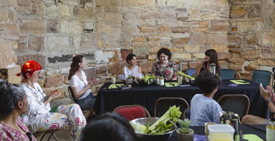 Six people are participating in a workshop together. They are gathered around a black rectangular table. There are various silver bowls and plates that are holding celery and fresh vegetables on the table.