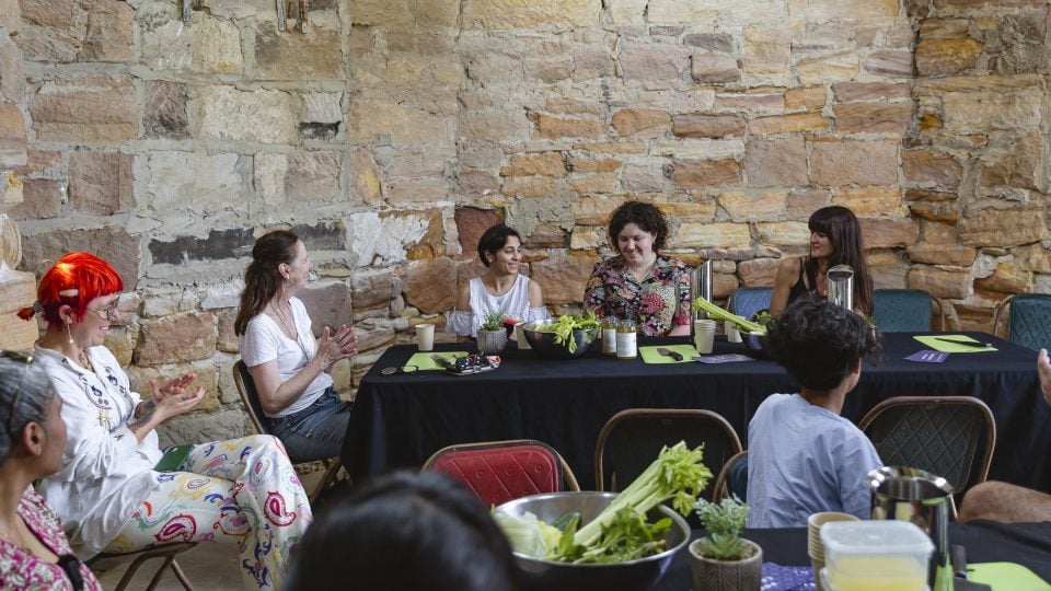 Six people are participating in a workshop together. They are gathered around a black rectangular table. There are various silver bowls and plates that are holding celery and fresh vegetables on the table.