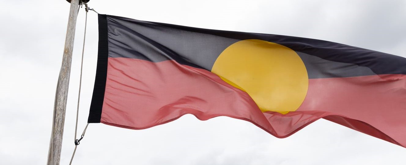 The Aboriginal flag flying in the wind on a post. There is a grey overcast in the sky behind the Aboriginal flag.