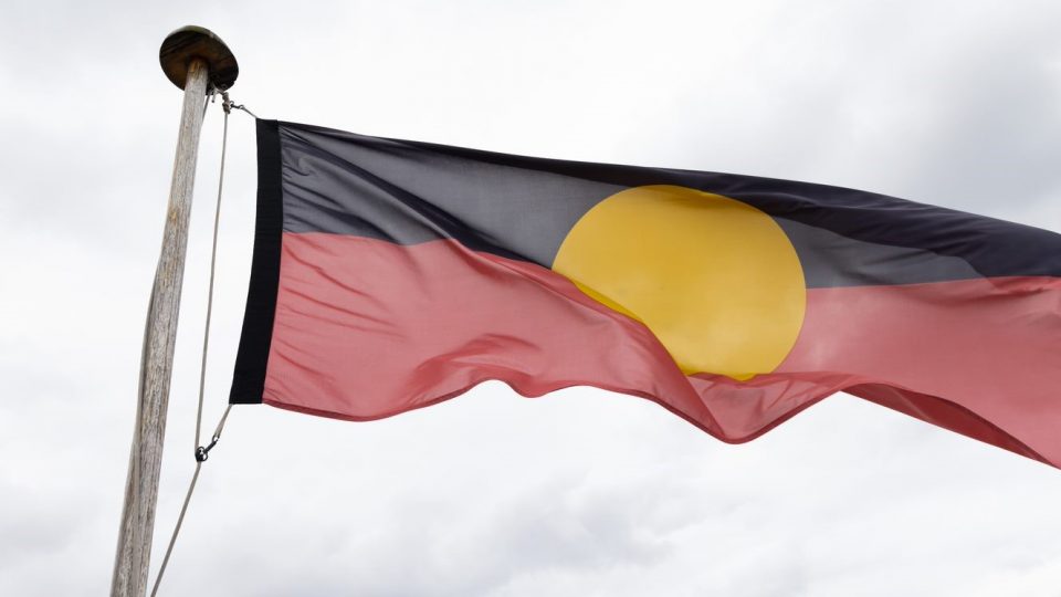 The Aboriginal flag flying in the wind on a post. There is a grey overcast in the sky behind the Aboriginal flag.