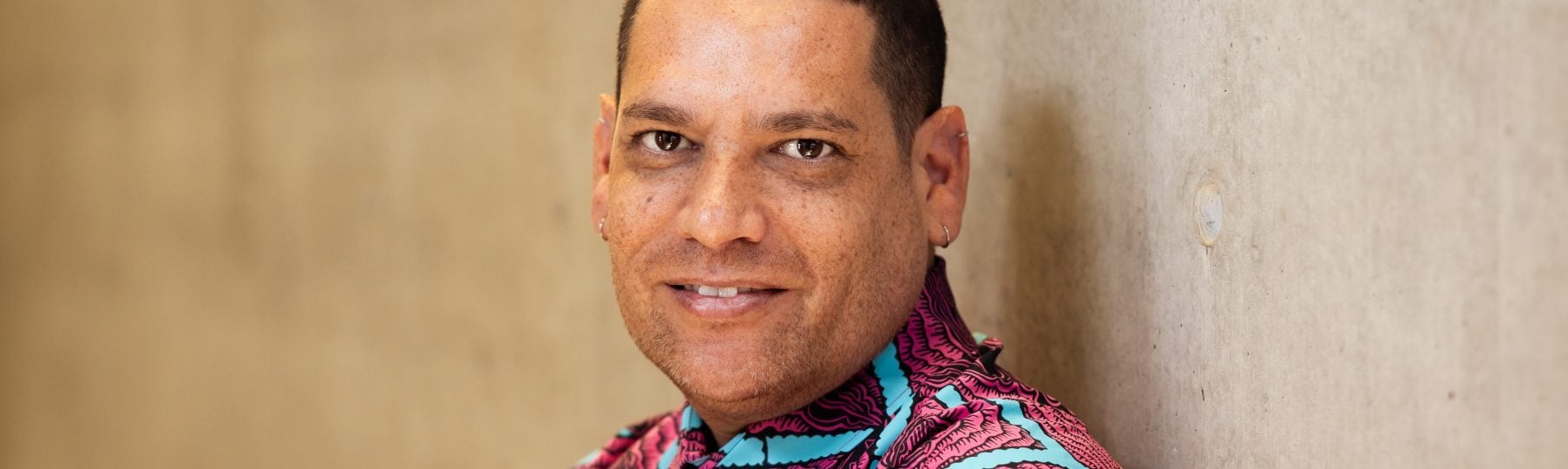 Tony Albert has arms crossed, with his back leaning against the wall. He is smiling and appears to be relaxed. He is wearing a pink and blue button up shirt.