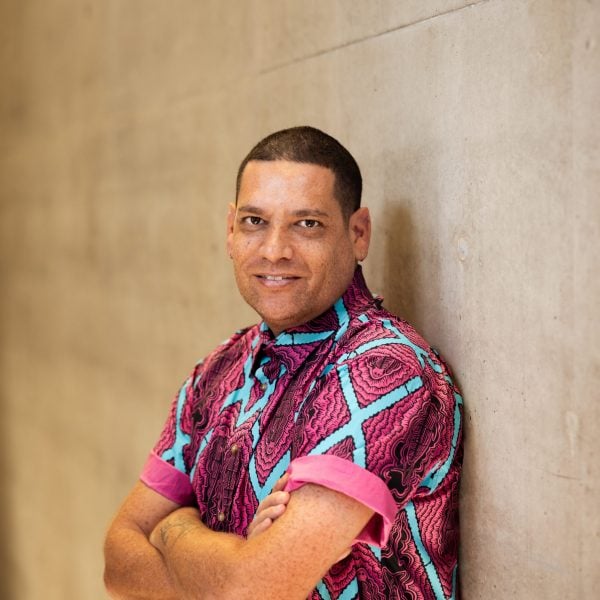 Tony Albert has arms crossed, with his back leaning against the wall. He is smiling and appears to be relaxed. He is wearing a pink and blue button up shirt.