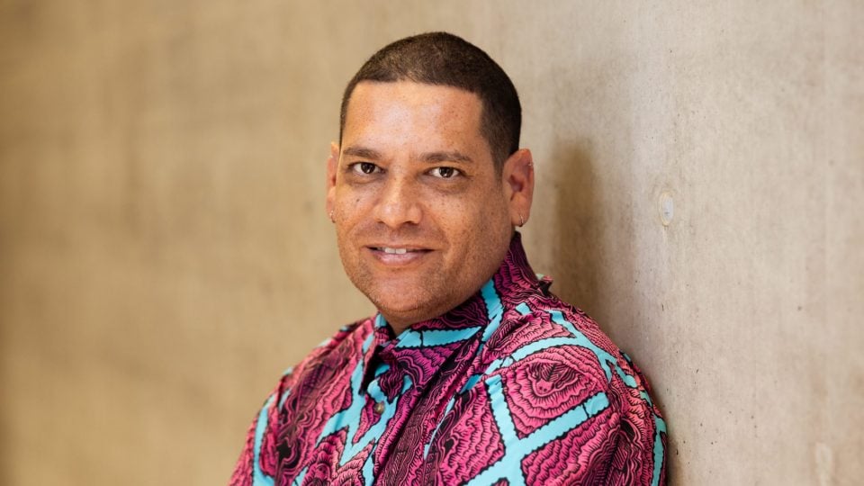 Tony Albert has arms crossed, with his back leaning against the wall. He is smiling and appears to be relaxed. He is wearing a pink and blue button up shirt.