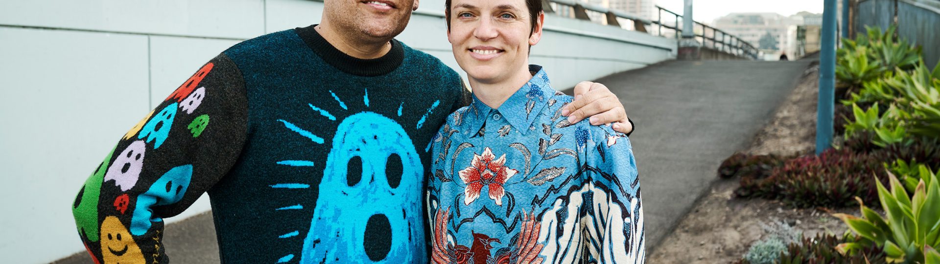 Tony Albert and Vivian Ziherl stand next to each other. Tony's arm is around Vivian's shoulder. Tony is wearing a black jumper with a large blue ghost on the front. Vivian is wearing a silk batik with floral arrangements and an eagle on it. They are both smiling.