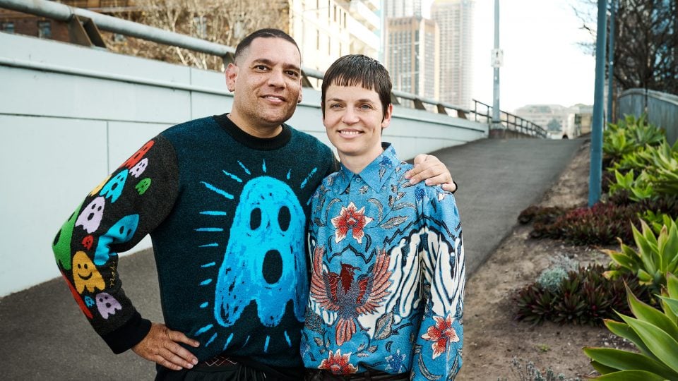 Tony Albert and Vivian Ziherl stand next to each other. Tony's arm is around Vivian's shoulder. Tony is wearing a black jumper with a large blue ghost on the front. Vivian is wearing a silk batik with floral arrangements and an eagle on it. They are both smiling.