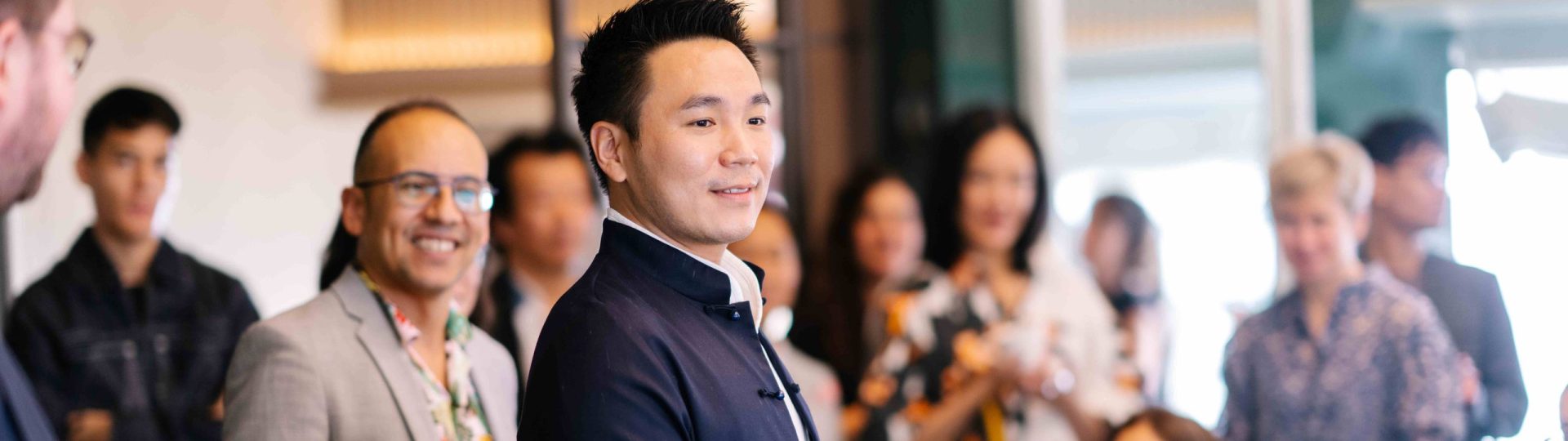 An Asian man is dressed in a fitted dark blue suit. He is delivering a speech as he stands up in front of a crowd.