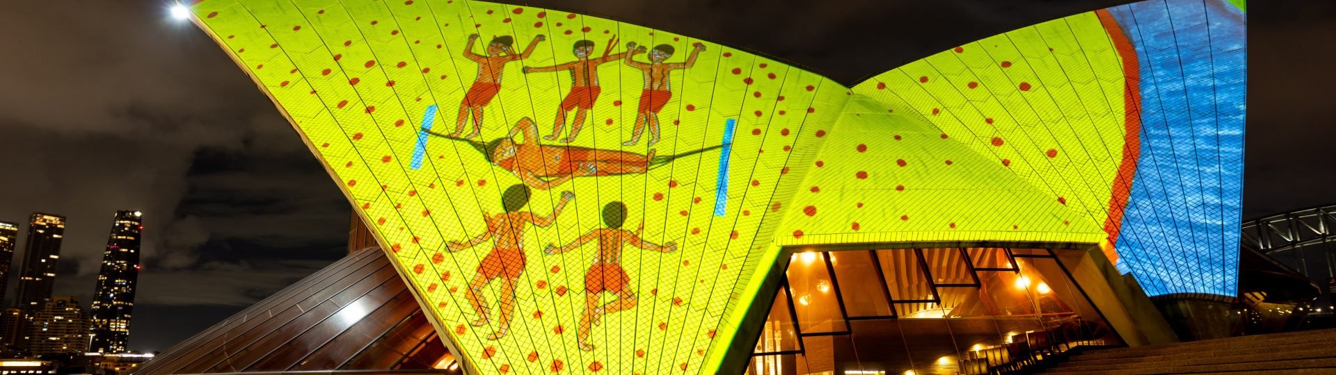 Dynamic projections Badu Gili displaying drawings by Joseca Mokahesi Yanomami on the eastern Bennelong sails of the Sydney Opera House. The drawing is a group of four surrounding a person in a hammock, sand is represented by a large yellow background with red dots, on the right of the drawing the sea is established in blue.